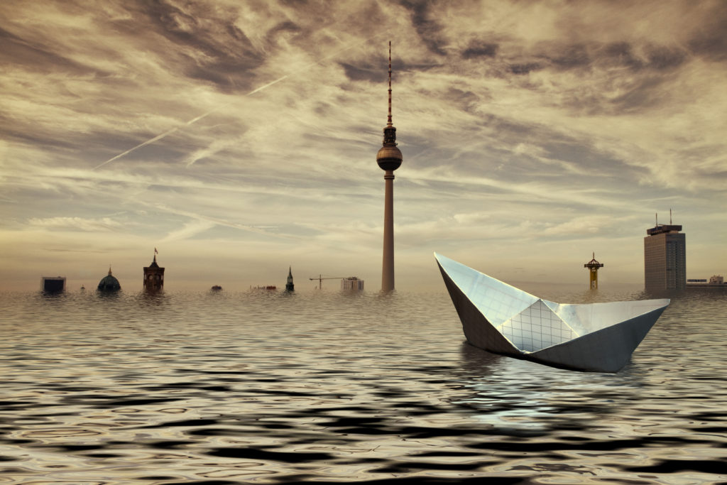 Paper boat adrift in the waters of a flooded Berlin, against the skyline of the city with the TV Tower (Fernsehturm) and the Park Inn Hotel, the Rotes Rathaus (Red City Hall) and the dome of the Berliner Dom (Berlin Cathedral) in the background.